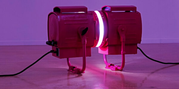 Twee grote, industriële, roze lampen staan op de grond recht tegenover en vlakbij elkaar en schijnen in elkaars richting, in een witte exporuimte die baadt in paars licht. 

Two large, industrial, pink lamps stand right in front of each other, facing each other, shining their light into each other, in a white exhibition space bathing in purple light.