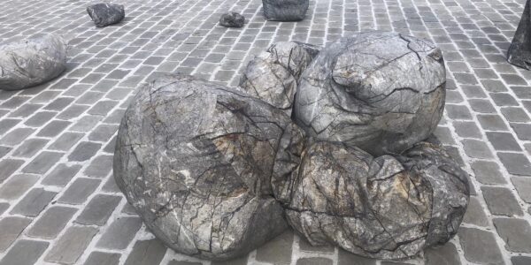 Steenachtige vormen ter grootte van een mens liggen op straat, op kasseien naast het water. 

Rock-like shapes the size of a person lie on the street, on a cobbled are next to a bassin of water.