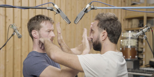 Twee mannen staan recht tegenover elkaar in een geluidsstudio, met in totaal 4 microfoons boven en achter hen, en tikken of slaan elkaar in het gezicht. 

Two man stand facing each other in a sound studio, with a total of 4 microphones above and behind them, while slapping each other slightly in the face.