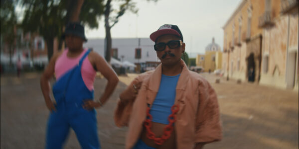 Twee mensen staat in een zonnige straat, een persoon scherp en een persoon wazig, ze kijken allebei in de camera en dragen pastelkleurige kledij in tinten blauw en roze. 

Two people in a sunny street setting, one in focus and one blurry, looking at the camera, wearing pastel coloured clothing in shades of blue, salmon and pink.