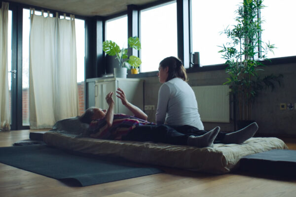 Vrouw ligt op haar rug op een matras op de grond, in een lichte ruimte met planten, en vertelt iets aan een andere vrouw die naast haar op de grond zit. 

Woman lies on her back on a mattress on the floor, in a light room with plants, and tells something to another woman sitting next to her on the floor.