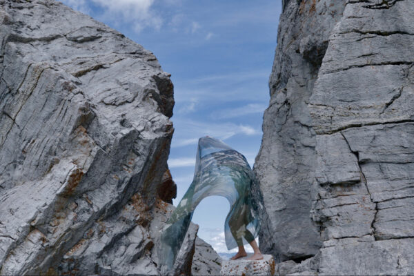 Rotsachtig landschap met blauwe lucht, met in het midden een persoon op blote voeten op een stenen pilaar, met een groen transparant doek over zich. 

Rocky landscape with blue sky, with a person in the middle, standing barefoot on a stone pillar, covered by a green transparent piece of cloth.