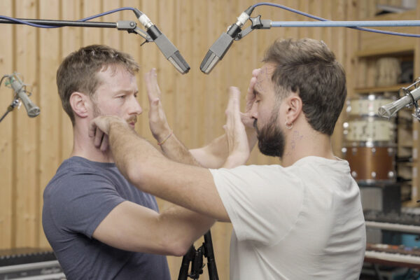 Twee mannen staan recht tegenover elkaar in een geluidsstudio, met in totaal 4 microfoons boven en achter hen, en tikken of slaan elkaar in het gezicht. 

Two man stand facing each other in a sound studio, with a total of 4 microphones above and behind them, while slapping each other slightly in the face.