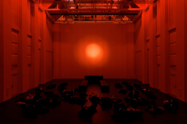 Een groep mensen ligt op de grond tussen enkele subwoofers, in een grote, witte, industriële ruimte die baadt in rood licht. 

A group of people lies on the floor between subwoofers, in a huge white industrial room bathing in red light.