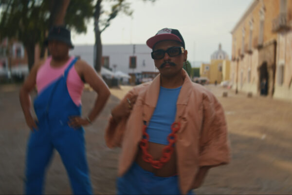 Twee mensen staat in een zonnige straat, een persoon scherp en een persoon wazig, ze kijken allebei in de camera en dragen pastelkleurige kledij in tinten blauw en roze. 

Two people in a sunny street setting, one in focus and one blurry, looking at the camera, wearing pastel coloured clothing in shades of blue, salmon and pink.