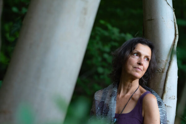 Vrouw met een paars topje staat in een bos, haar rug leunend tegen een witte boom, en kijkt dromerig naar links. 

Woman wearing a purple top stands in a forest, leaning her back on a white tree, looking dreamily to her left.
