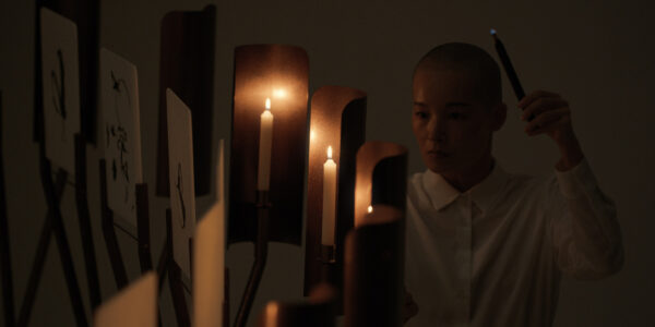 Persoon met kaal hoofd houdt een kaars vast bij kaarslicht, weerspiegeld in spiegels tussen tekeningen, met een stille, rituele sfeer.

Person with shaved head holds a candle by candlelight, reflected in mirrors among drawings, creating a quiet, ritual-like atmosphere.