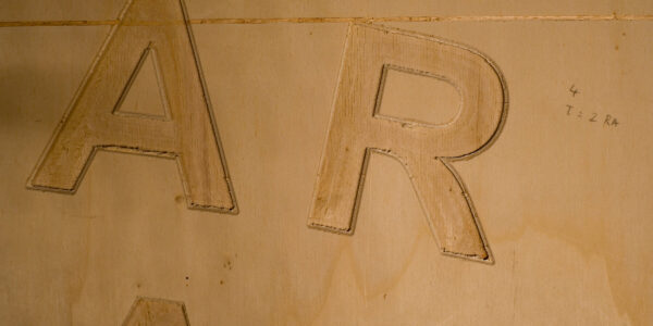 Houten plank waaruit grote letters A en R gefreesd zijn, met bovenaan een smalle gefreesde horizontale lijn die door de top van de A loopt. 

Wooden plank showing large letters A and R milled out, with a narrow horizontal milled out part crossing the top part of the A.