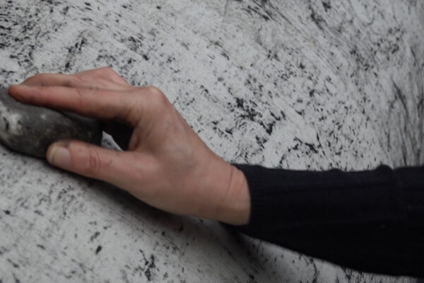 Een hand wrijft met een steen op de schors van een boom die omwikkeld is met witte stof, waardoor tekeningen ontstaan op de stof. 

A hand rubs a stone on the bark of a tree that is covered in white cloth, creating marks on the cloth.